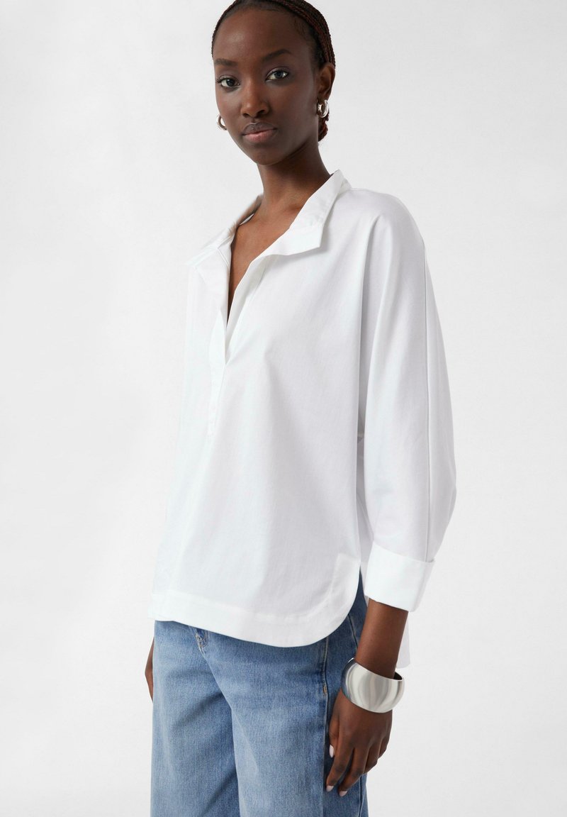 White collared blouse with a relaxed fit, long sleeves, and a rounded hem. Paired with light blue jeans and a wide metallic bracelet.