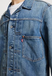 Džínová bunda světle modré barvy s vertikálními pruhy. Obsahuje stříbrné knoflíky, dvě p�řední kapsy a červený logový štítek na levé kapse.