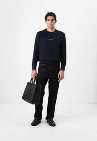 Sweater azul-marinho com logótipo branco, jeans pretos e mala de couro preta com design estruturado. Modelo segura a mala pelas alças. Sapatos pretos.