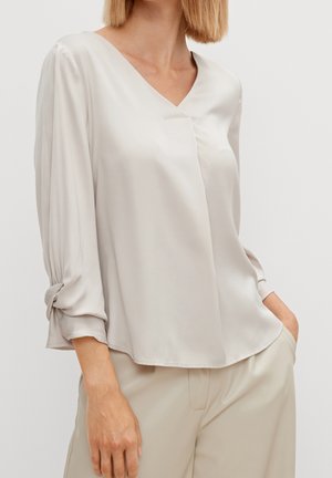 Femme portant un chemisier en satin beige clair à col en V avec manches 3/4 et pantalon beige, une main dans la poche sur fond uni.