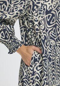 Navy-blaues Kleid mit abstraktem beigem Muster, mit elastischen Bündchen und Seitentasche. Hergestellt aus Textilgewebe mit lockerer Passform.