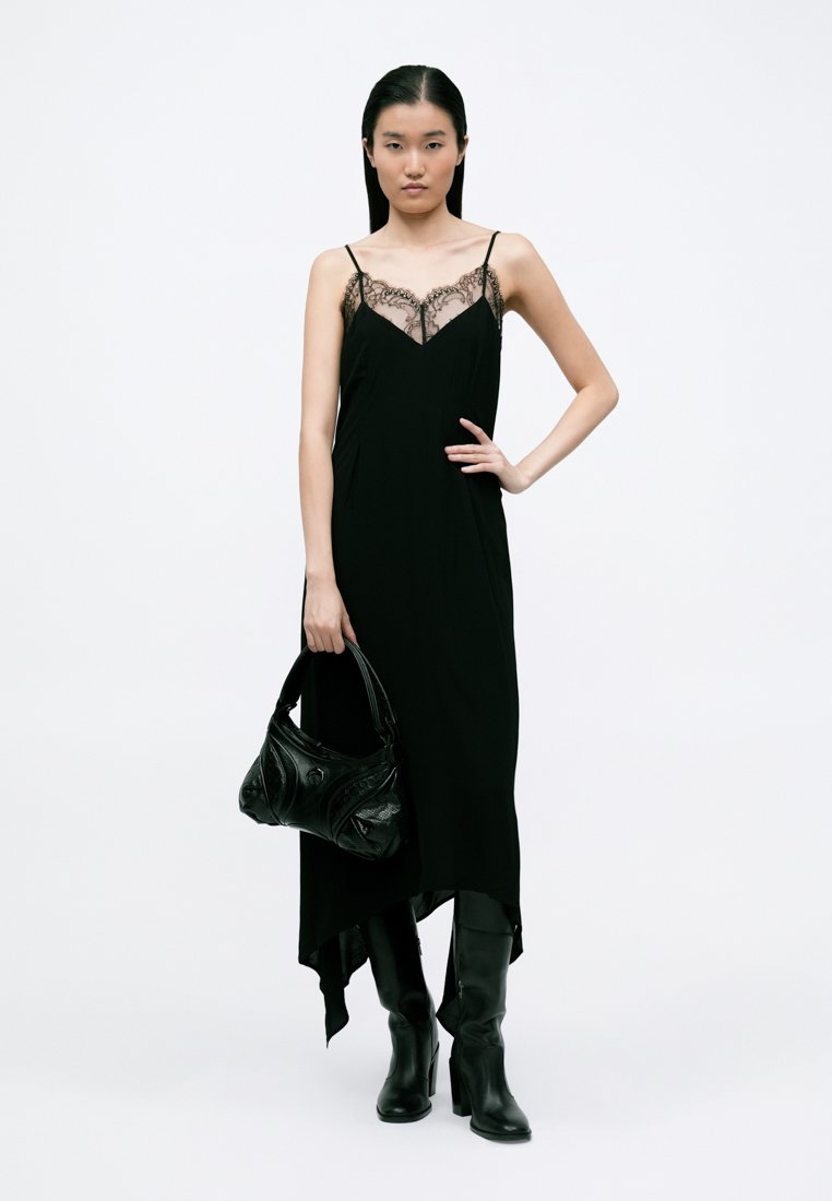 Black dress with lace trim, spaghetti straps, and asymmetrical hem, paired with a textured black handbag and knee-high leather boots.