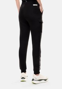 Pantaloni jogger neri con design affusolato, dotati di tasche cargo laterali, dettagli lucidi e polsini elasticizzati, abbinati a scarpe da ginnastica bianche.