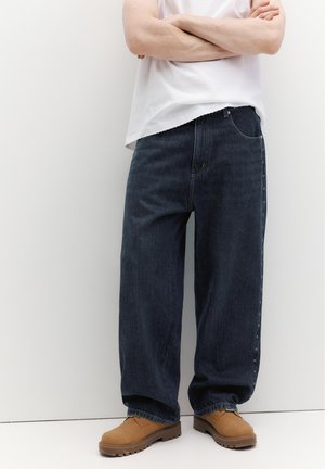 Személy laza, sötétkék farmernadrágot visel, oldalán csillagmintákkal, barna fűzős bakancsot és fehér rövid ujjú felsőt, karjait összefonta.