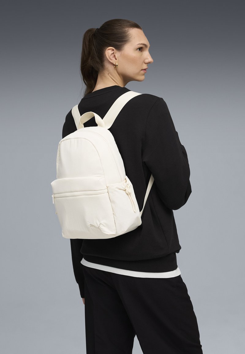 Sac à dos crème avec une forme arrondie, comportant une poche zippée à l'avant, des bretelles réglables et une finition en tissu texturé.