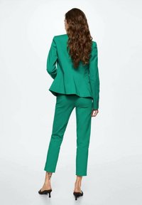 Costume vert ajusté avec un blazer structuré et un pantalon slim-fit. Le tissu a une texture lisse, avec un détail peplum et un design minimaliste.