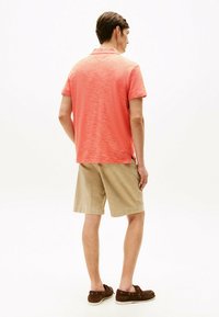 Polo a maniche corte di colore corallo con un motivo testurizzato, abbinato a pantaloni corti color khaki. Modello che indossa scarpe da barca marroni, visto da dietro.