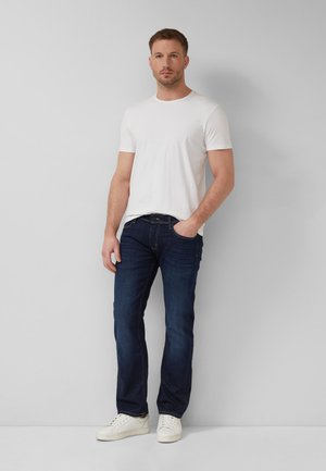 Man staat tegen een grijze muur en draagt een wit T-shirt met korte mouwen, donkerblauwe spijkerbroek, witte sneakers, met één hand in de zak.