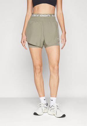 Kvinde iført olivengrønne lagdelte sportsshorts, sort cropped top, hvide Nike-strømper og hvide løbesko, stående foran en ensfarvet baggrund.