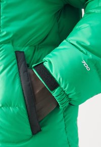 Zelená hřejivá bunda s prošívanou texturou, černými suchými zipy a číslem "700" vytištěným na rukávu. Detailní pohled na manžetu.