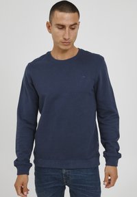 Navy blue sweatshirt made of cotton, featuring a crew neck, ribbed cuffs and hem, with a small logo on the chest. Worn with denim jeans.