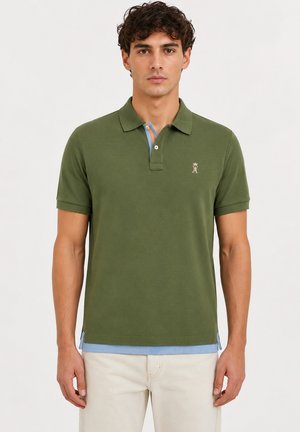Jeune homme aux cheveux courts et bouclés porte un polo vert olive par-dessus un sous-pull bleu clair et un pantalon beige, debout devant un fond uni.