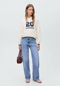 Cream sweater with navy print "20 CLASSICS," paired with light blue wide-leg jeans. Model holds a burgundy handbag, wearing light pink shoes.