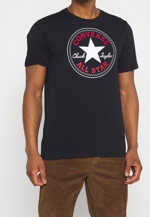 Homme portant un t-shirt noir avec une étoile blanche et un logo rouge "Converse All Star Chuck Taylor", associé à un pantalon marron.