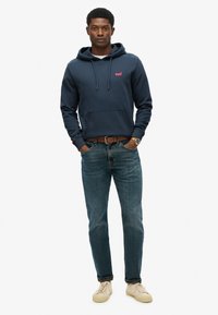 Marineblauwe hoodie met een voorzak, koordjes en een roze logo; gecombineerd met lichtblauwe jeans en beige sneakers.