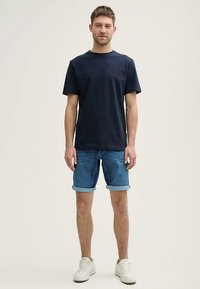 Marineblå bomulds-T-shirt med rullede ærmer; blå denimshorts med opsmøg; hvide sneakers. Modellen står afslappet på en neutral baggrund.