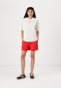 Chemise blanche en mesh à manches courtes et col en V, assortie à un short rouge. Le modèle porte des baskets noires avec des lacets beiges et une semelle en caoutchouc.