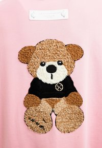 Tela rosa con un oso de peluche bordado en marrón y crema, que lleva una camiseta negra con un logo. Textura suave y costuras detalladas.