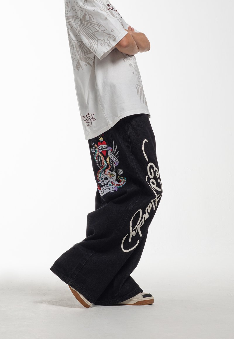 Zwarte denim wijde broek met kleurrijke borduursels van een schedel en een slang, en witte tekstaccenten aan de zijkant.