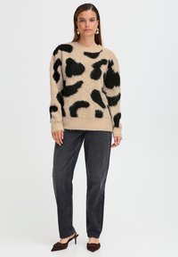 Pull en maille beige avec imprimé léopard noir, col rond, manches longues et tissu texturé ; associé à un jean droit gris foncé et des talons marron.