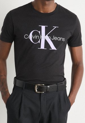 Sort kortærmet t-shirt lavet af bomuld, med "Calvin Klein Jeans" logo i hvid og lys lavendel. Teksten er centreret på fronten.