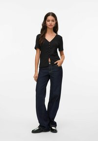 Svart kortärmad V-ringad topp med knäppning fram, kombinerat med mörkblå vida jeans och svarta skor med metalliska detaljer.