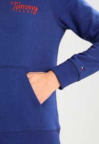 Felpa in cotone blu navy con tasca frontale, polsini a costine e logo "Tommy Jeans" ricamato in rosso sul petto.