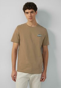 Junger Mann, der ein beiges T-Shirt mit einem kleinen gestickten Oldtimer auf der Brust trägt, kombiniert mit einer hellen Hose, steht vor einer grauen Wand.