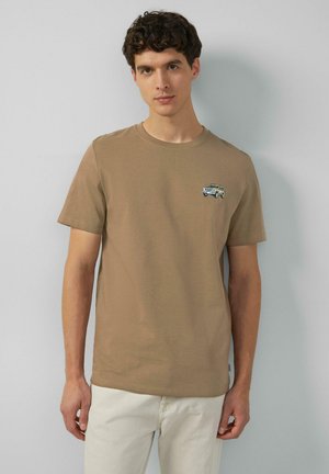 Junger Mann, der ein beiges T-Shirt mit einem kleinen gestickten Oldtimer auf der Brust trägt, kombiniert mit einer hellen Hose, steht vor einer grauen Wand.