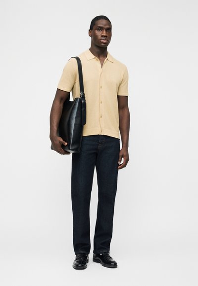 Άνδρας που φοράει μπεζ κοντομάνικο πουκάμισο με κουμπιά, σκούρο τζιν, μαύρα παπούτσια, κρατώντας μια μεγάλη μαύρη δερμάτινη τσάντα ώμου σε ένα χέρι.
