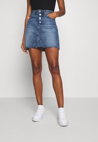 Jeans A-linje kjol med femknappsstängning, fransad nederkant och sidofickor; medelblå tvätt; kombinerad med vita skor och en svart topp.