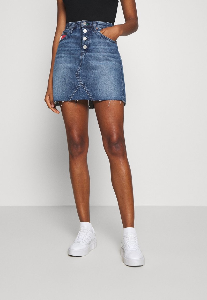 Jeans A-linje kjol med femknappsstängning, fransad nederkant och sidofickor; medelblå tvätt; kombinerad med vita skor och en svart topp.