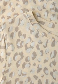 Beżowa tkanina z wzorem w cętki pantery w odcieniach jasnobrązowym i srebrnym, ukazująca zbliżenie szwów przy dekolcie i rękawie.