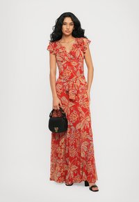 Robe maxi fleurie rouge avec des manches courtes à volants, un détail de taille nouée et une jupe fluide. Le mannequin tient un sac à main noir.