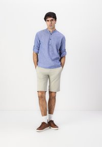 Lys blå, knappet langermet skjorte med oppbrettede ermer, kombinert med beige shorts. Brune sneakers og hvite sokker fullfører antrekket.