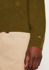 Olivově zelený pletený svetr s žebrovanou strukturou, na hrudi s malým logem a pruhovaným akcentem na manžetě. Spárován s béžovými kalhotami.