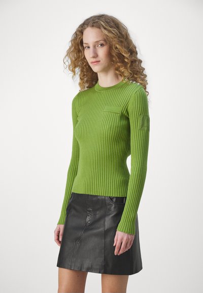 Grüner gerippter Strickpullover mit Rundhalsausschnitt, der eine Tasche an der Vorderseite und silberne Akzente an der Schulter aufweist, kombiniert mit einem schwarzen Lederrock.