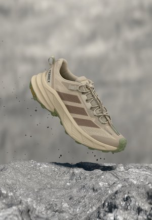 Beige Adidas Terrex vandresko med grøn ydersål, svævende over stenfuldt terræn, viser sideprofil og detaljeret mesh-overdel.