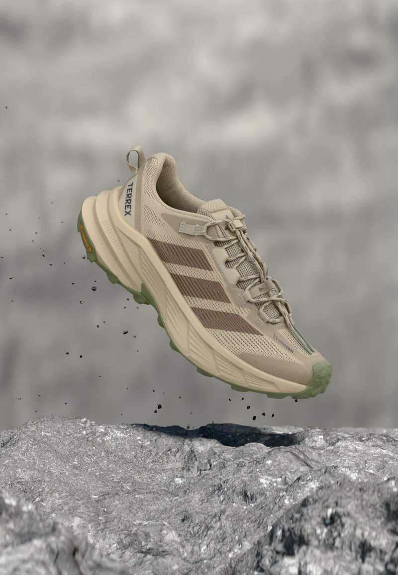 Бежова туристическа обувка Adidas Terrex с зелена подметка, във въздуха над rocky терен, показваща страничен изглед и детайлирана горна част от мрежест материал.