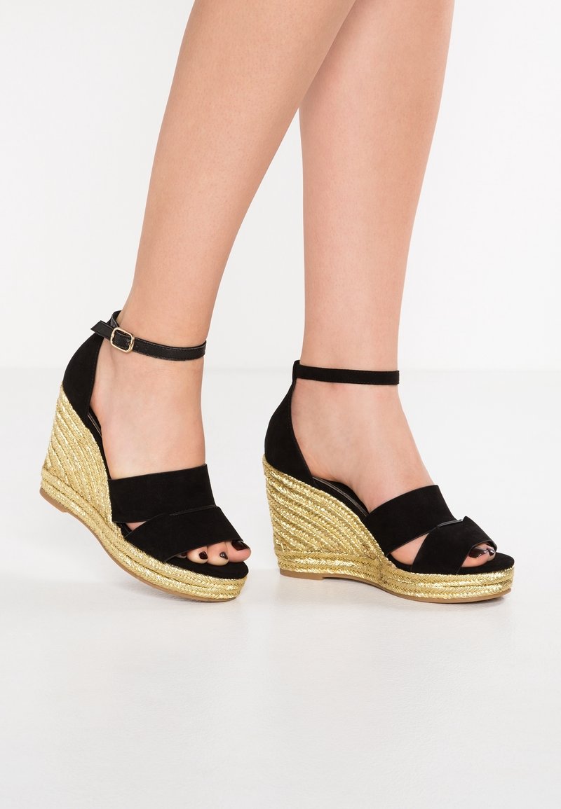 Jambes portant des sandales compensées en daim noir avec des talons plateforme tressés dorés et des brides à la cheville sur fond blanc.