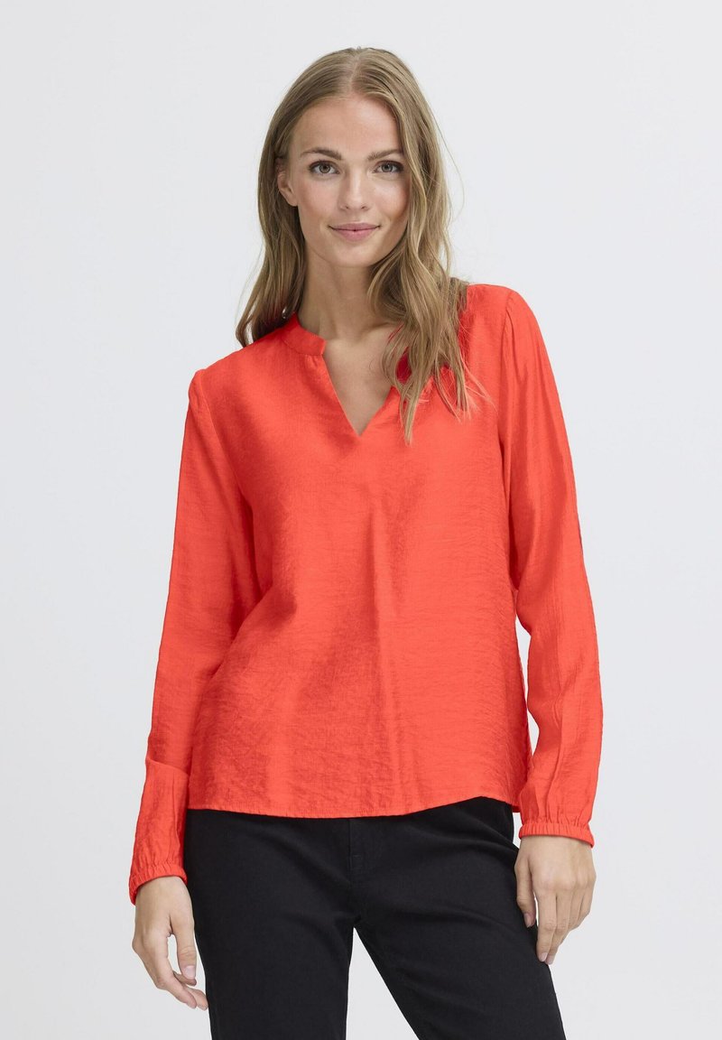 Rode blouse met lange mouwen en V-hals, zachte textuur en plooien bij de manchetten, gecombineerd met donkere broek.