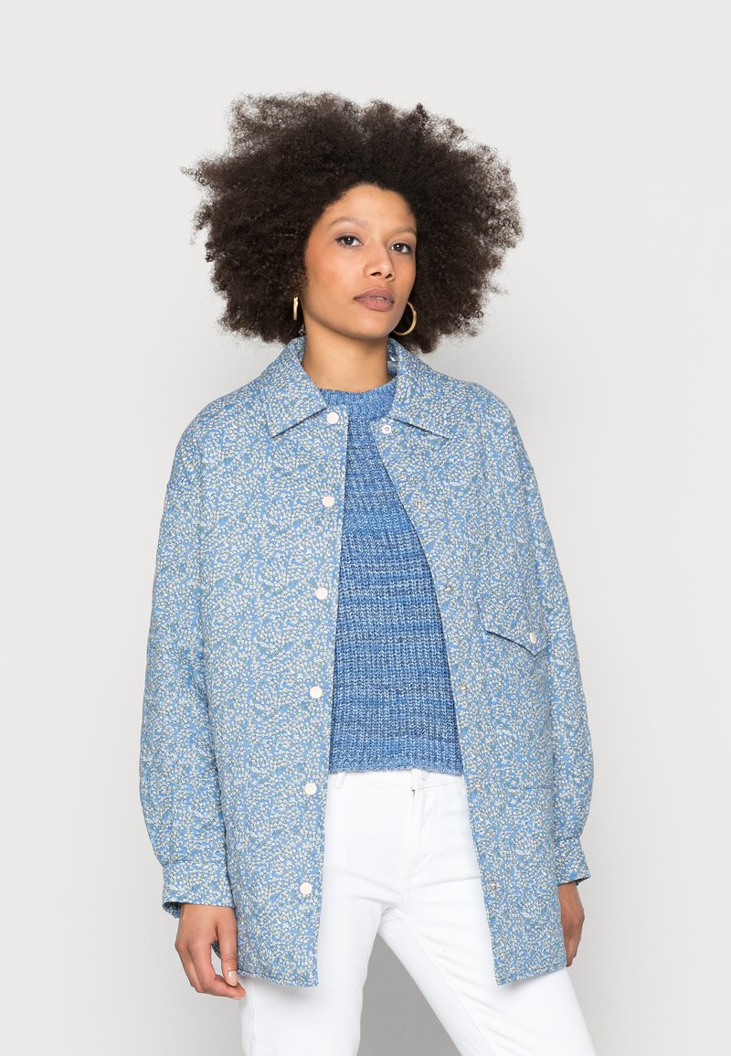Blue floral-patterned denim jacket with a relaxed fit, featuring a collared neck, snap buttons, and long sleeves, worn over a textured blue sweater.