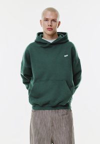 Junge Person mit kurzen blonden Haaren, die einen dunkelgrünen Kapuzenpullover und hellbraune Hosen trägt, steht mit den Händen hinter dem Rücken.