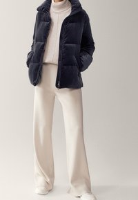 Kobieta w granatowej pikowanej kurtce, jasnobeżowym golfie, kremowych spodniach o szerokich nogawkach i białych trampkach, stojąca z rękami w kieszeniach.