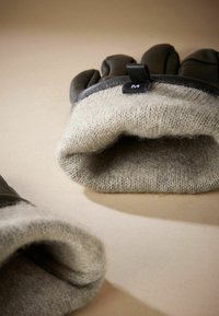 Läderhandskar med ett varmt, ljusgrått stickat foder. Funktioner inkluderar en slät yta, vikbar manschett och en svart etikett märkt "M."