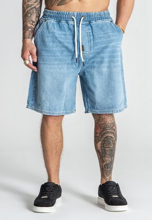 Shorts en denim bleu clair avec une taille élastique, fermeture à cordon, deux poches latérales et une coupe décontractée. Portés avec des baskets noires.