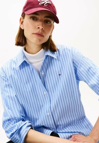 Camicia a righe blu chiaro con dettagli rossi, con colletto e bottoni, indossata sopra un dolcevita bianco, abbinata a un berretto bordeaux.