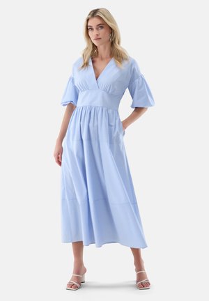 Blonde Frau, die ein hellblau gestreiftes Midikleid mit Puffärmeln und weißen Sandalen mit Absatz trägt, steht mit einer Hand in der Tasche.