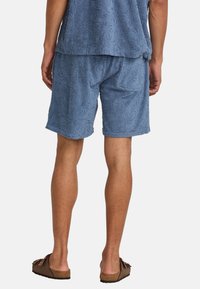 Blue toweling shorts with textured palm leaf pattern, elastic waistband, and side pockets. Worn with brown slip-on sandals.