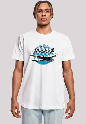 Junger Mann mit geflochtenem Haar, der ein weißes T-Shirt mit einem Grafikdruck eines Vintage-Pan-Am-Airways-Flugzeugs und hellblaue Jeans trägt, vor einfachem Hintergrund.